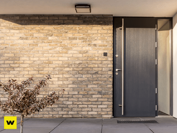 Moderne Haustür aus dunklem Holz mit Edelstahlgriff vor heller Klinkerfassade.